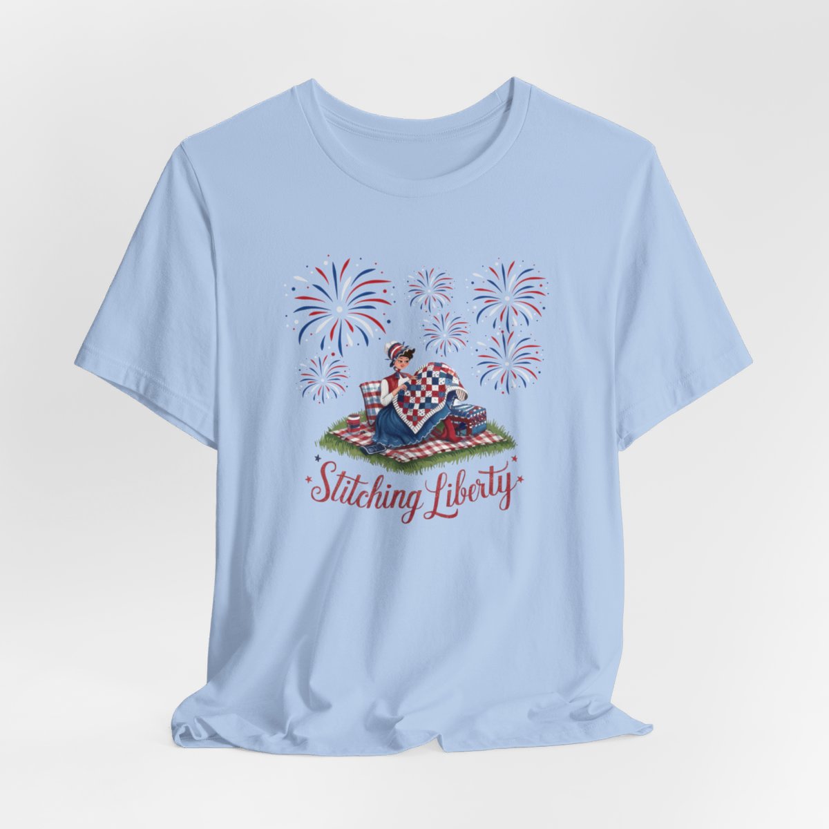 A Baby Blue T-shirt with illustrated scene of a colonial woman quilting a patriotic blanket, surrounded by fireworks. Text reads "Stitching Liberty" in a decorative font.