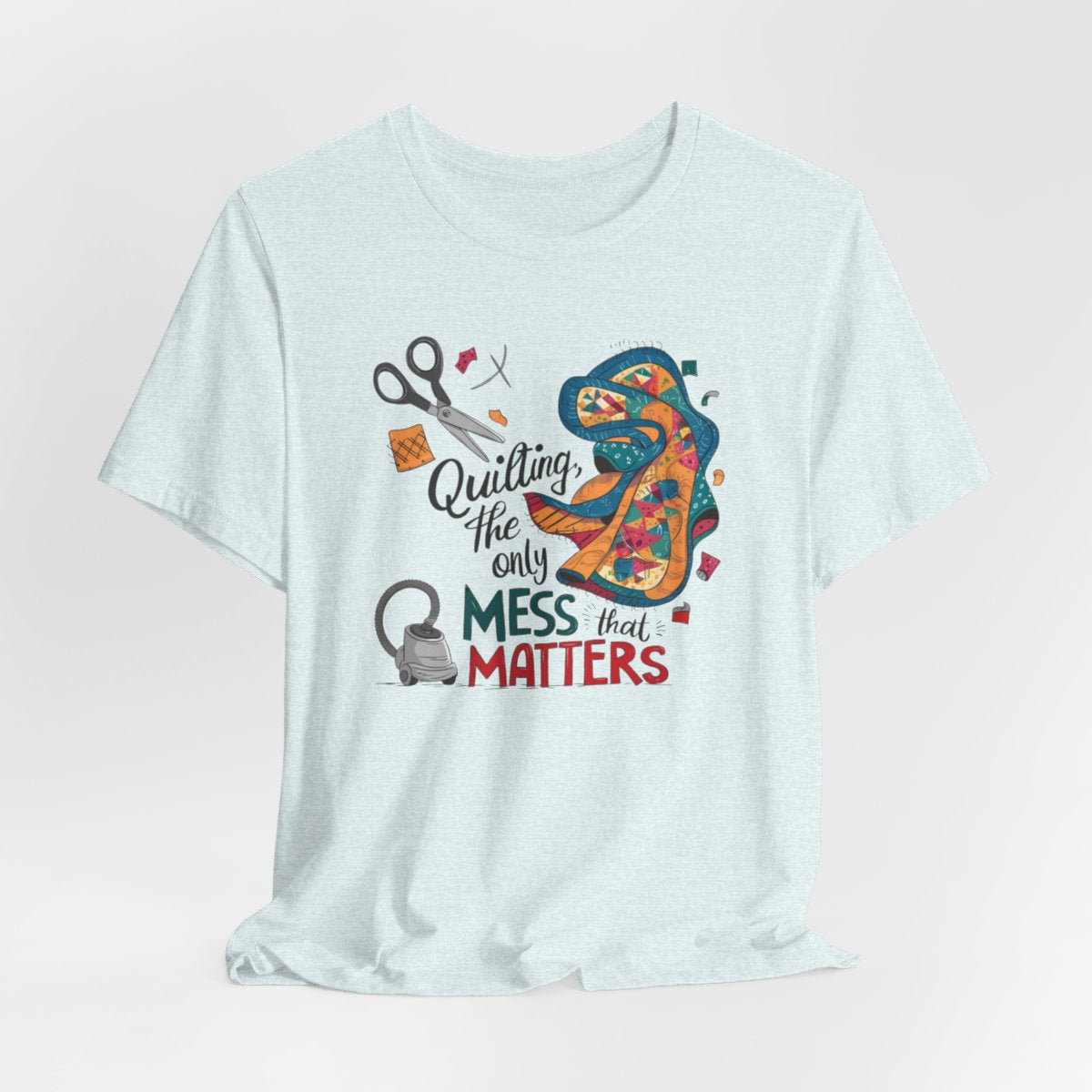 A Heather Ice Blue fun T-Shirt with the phrase Quilting, the Only Mess that Matters and illustrations of fabric scraps, scissors, and a vacuum cleaner. Colorful quilt patterns swirl around the text, creating a playful and relatable design for quilting enthusiasts.