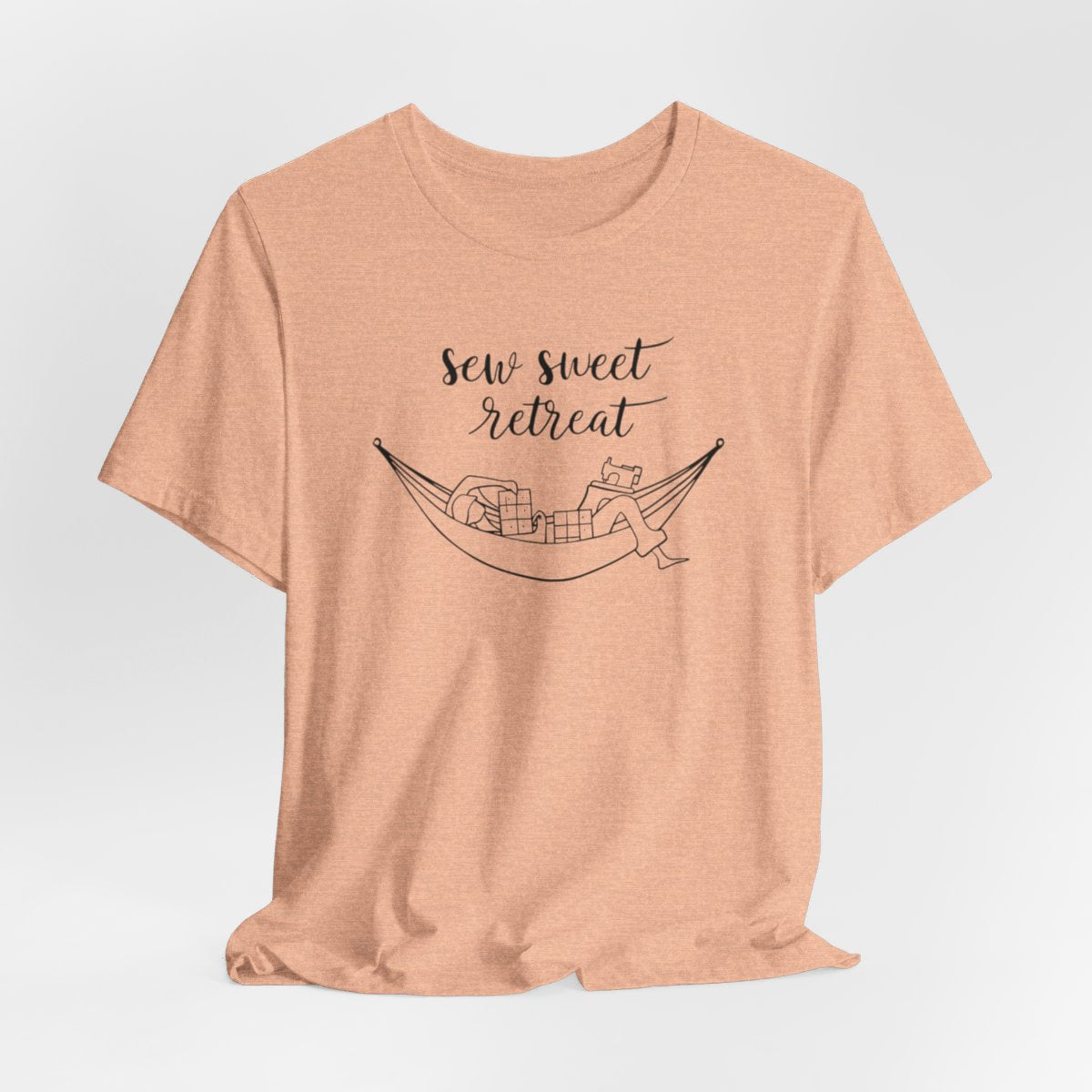 A Heather Peach T-Shirt with a black and white illustration of a hammock filled with quilting supplies like fabric squares and a sewing machine. Text above reads "Sew Sweet Retreat" in a flowing script font.