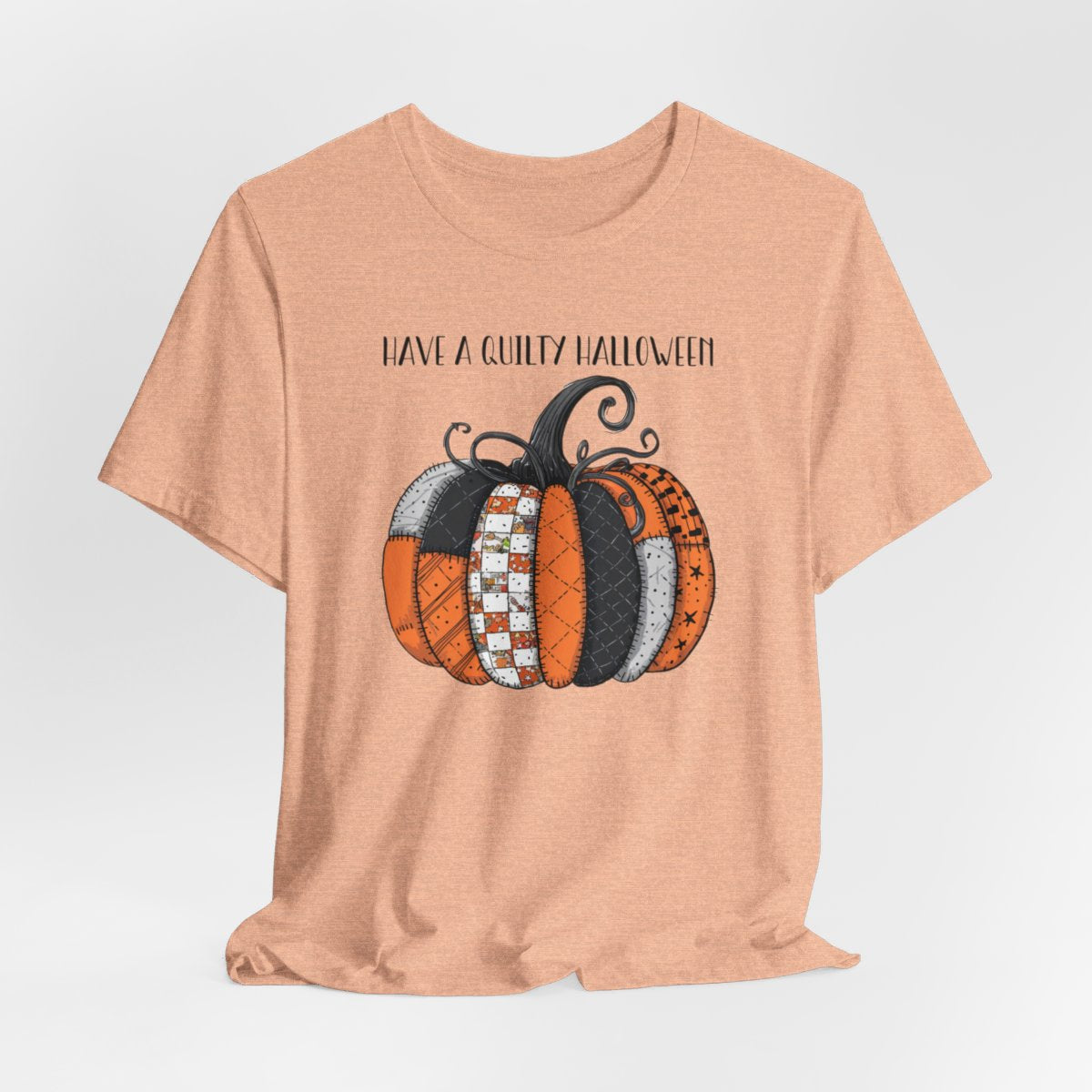 A Heather Peach Halloween Quilting T-Shirt featuring patchwork pumpkin with various quilt patterns. Mix of Halloween and traditional quilt motifs. Intricate stitching details. Text reads "Have a Quilty Halloween". Ideal for quilters who love Halloween.