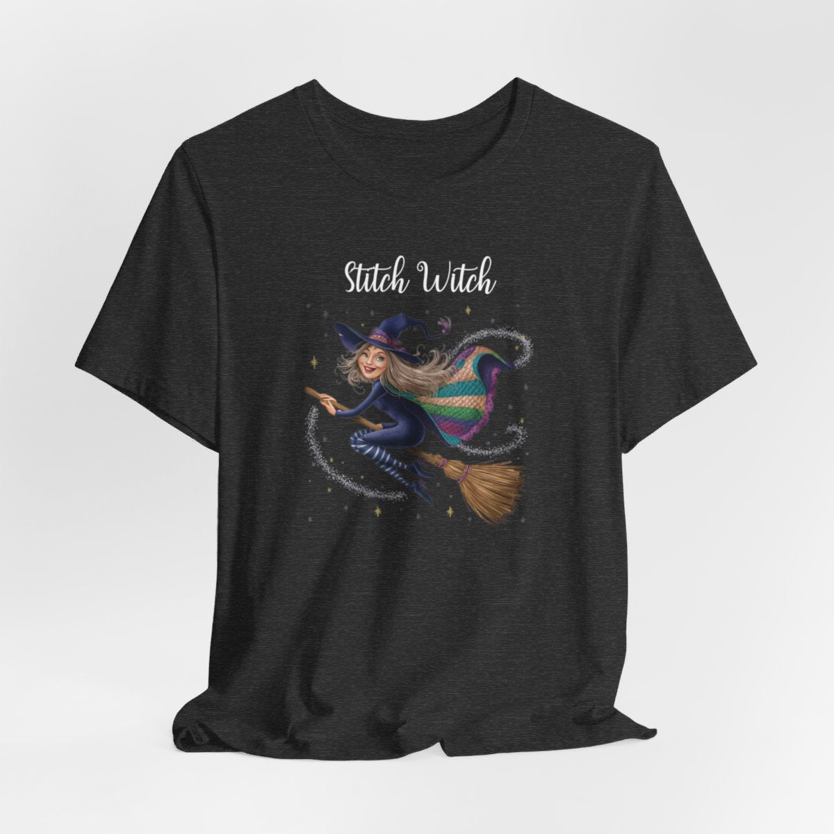 A Dark Grey Heather Halloween Quilting T-Shirt featuring a witch riding broomstick on t-shirt design. Colorful patchwork quilt cape with various patterns. Sparkling magic and stars. Text reads "Stitch Witch". Ideal for quilters who enjoy magical themes.