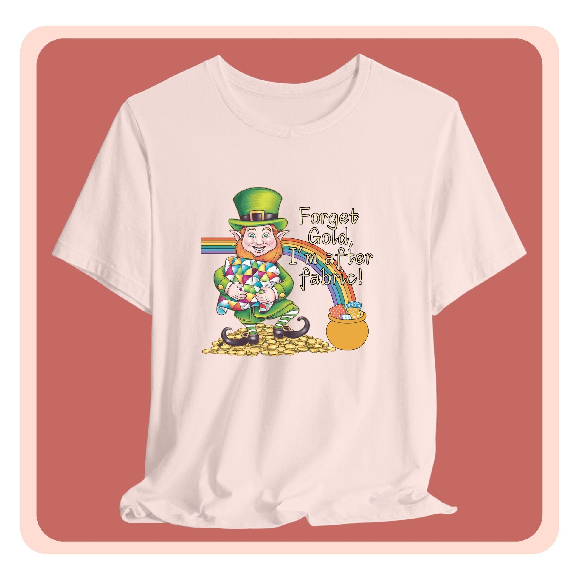 A short-sleeve t-shirt featuring a cheerful leprechaun holding a quilt, sitting on gold coins, with a rainbow leading to a fabric-filled pot and the phrase 'Forget Gold, I’m After Fabric!' Perfect for quilters celebrating St. Patrick’s Day.