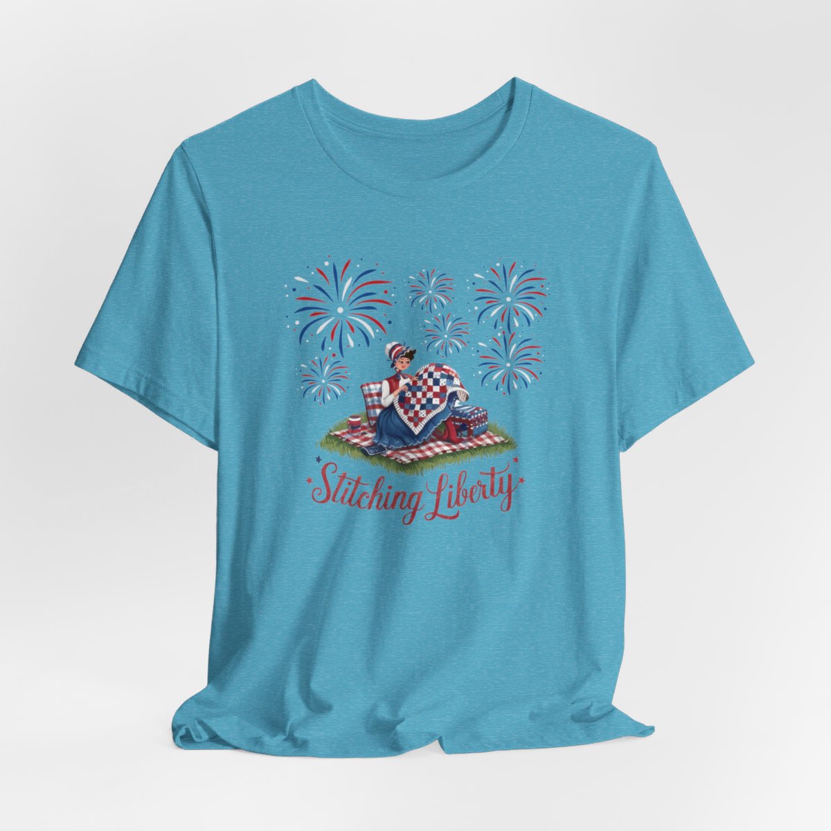 A Heather Aqua T-shirt with illustrated scene of a colonial woman quilting a patriotic blanket, surrounded by fireworks. Text reads "Stitching Liberty" in a decorative font.