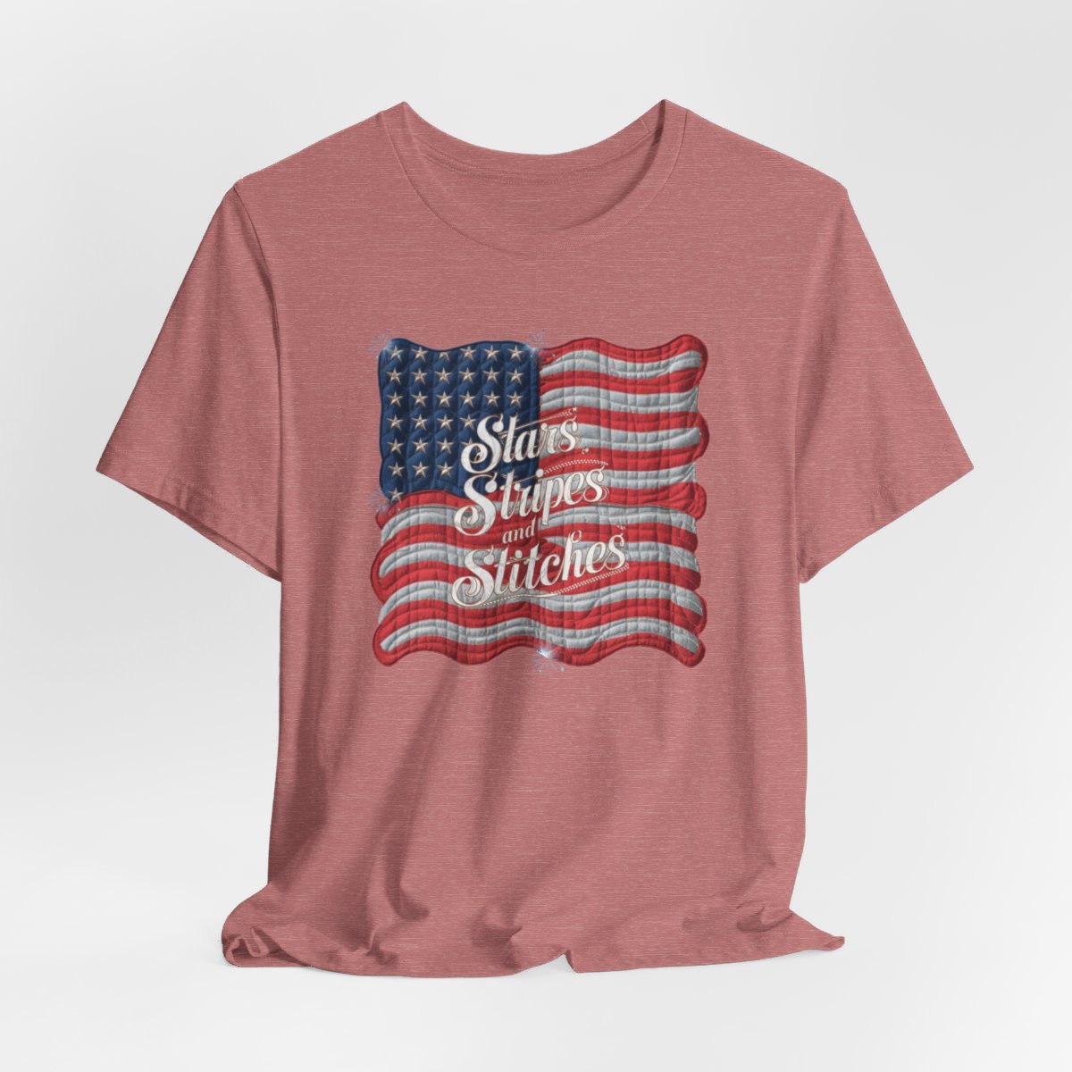 A Heather Mauve T-shirt featuring quilted American flag design with text "Stars Stripes and Stitches" overlaid in stylized, patriotic font. Fireworks accent the corners.