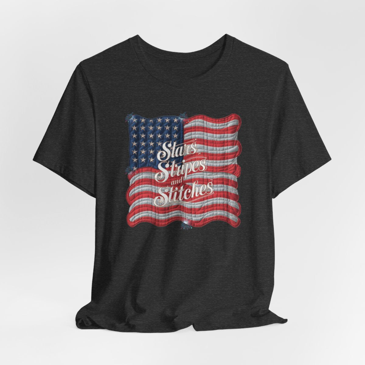 A Dark Grey Heather T-shirt featuring quilted American flag design with text "Stars Stripes and Stitches" overlaid in stylized, patriotic font. Fireworks accent the corners.