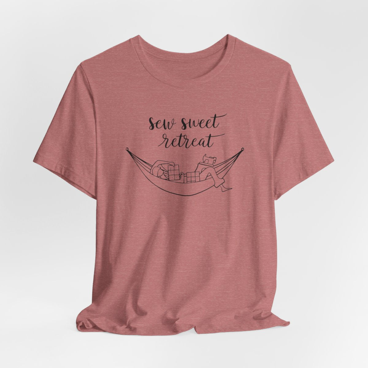 A Heather Mauve T-Shirt with a black and white illustration of a hammock filled with quilting supplies like fabric squares and a sewing machine. Text above reads "Sew Sweet Retreat" in a flowing script font.