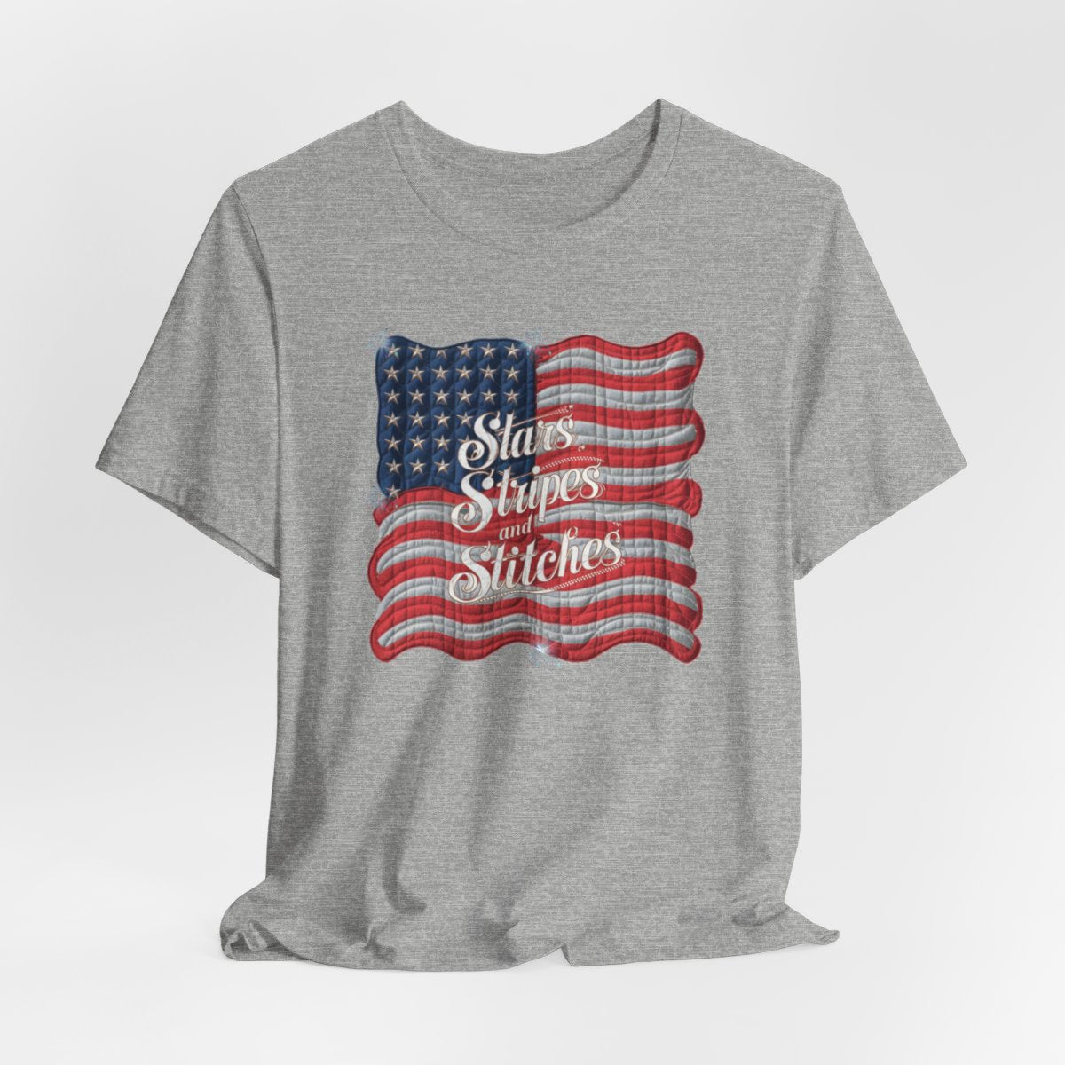 An Athletic Heather T-shirt featuring quilted American flag design with text "Stars Stripes and Stitches" overlaid in stylized, patriotic font. Fireworks accent the corners.