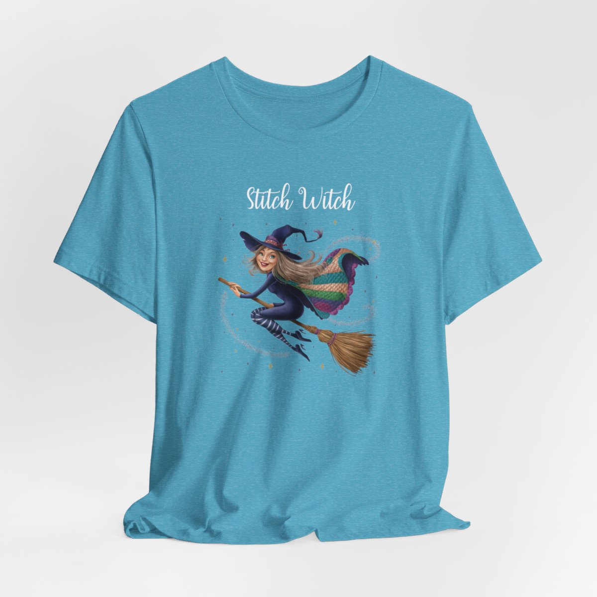 A Heather Aqua Halloween Quilting T-Shirt featuring a witch riding broomstick on t-shirt design. Colorful patchwork quilt cape with various patterns. Sparkling magic and stars. Text reads "Stitch Witch". Ideal for quilters who enjoy magical themes.