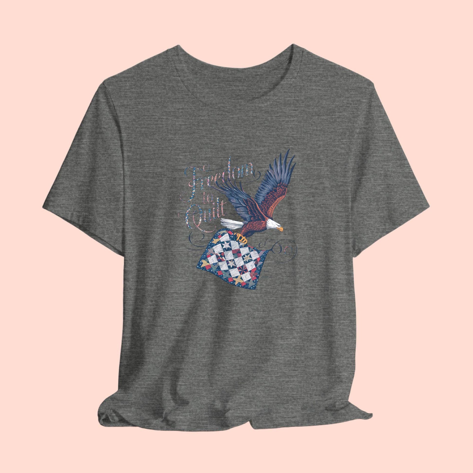 A T-shirt with detailed illustration of a bald eagle in flight, carrying a patchwork quilt. Text reads "Freedom to Quilt" in an ornate, patriotic font.