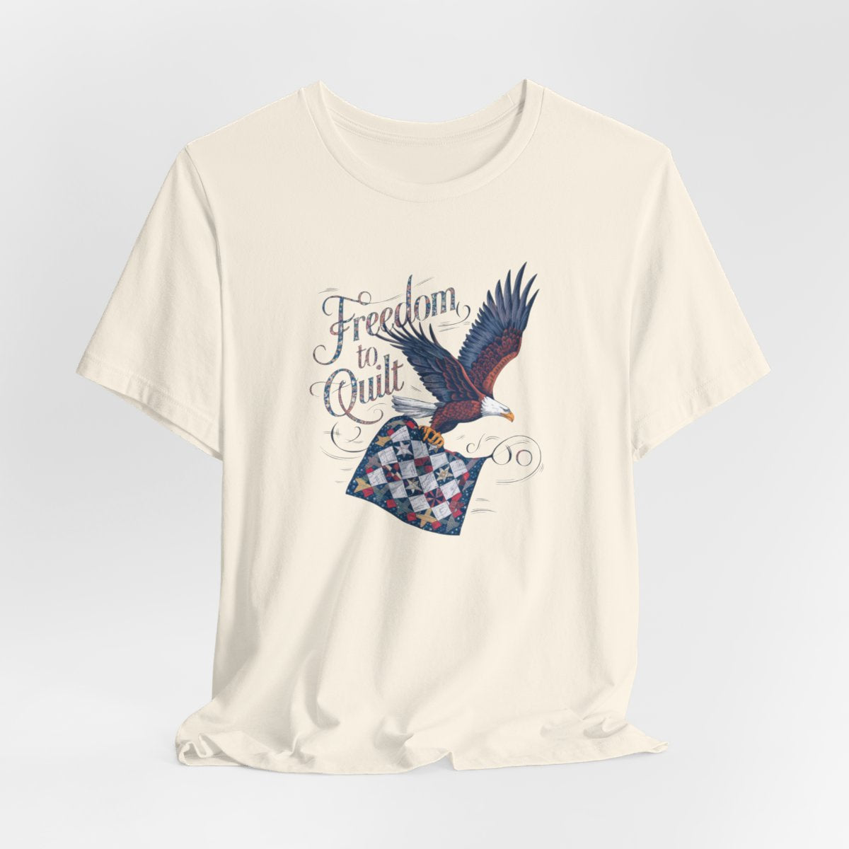 A Natural T-shirt with detailed illustration of a bald eagle in flight, carrying a patchwork quilt. Text reads "Freedom to Quilt" in an ornate, patriotic font.