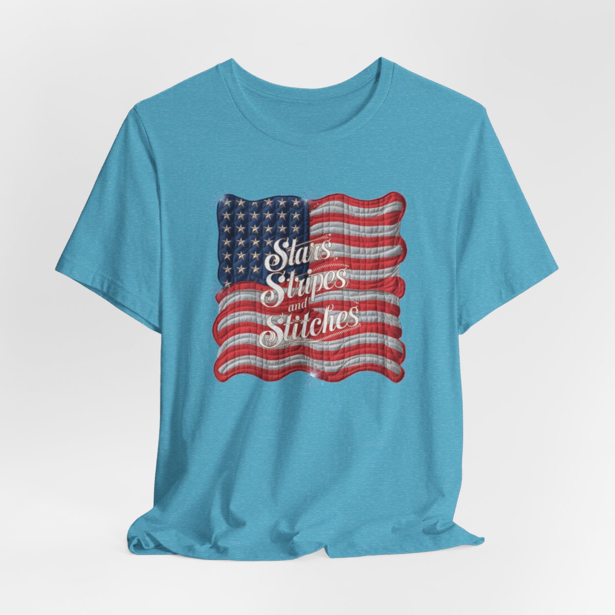 A Heather Aqua T-shirt featuring quilted American flag design with text "Stars Stripes and Stitches" overlaid in stylized, patriotic font. Fireworks accent the corners.