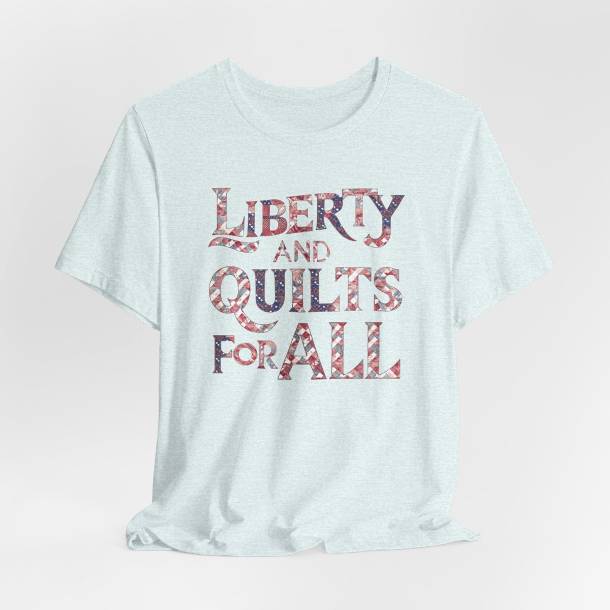 A Heather Ice Blue T-shirt featuring text "LIBERTY AND QUILTS FOR ALL" in large decorative letters filled with various quilt patterns in red, white, and blue.