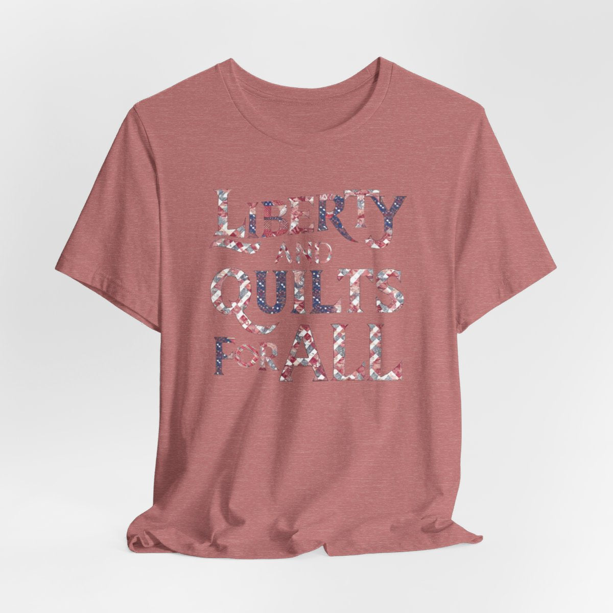 A Heather Mauve T-shirt featuring text "LIBERTY AND QUILTS FOR ALL" in large decorative letters filled with various quilt patterns in red, white, and blue.