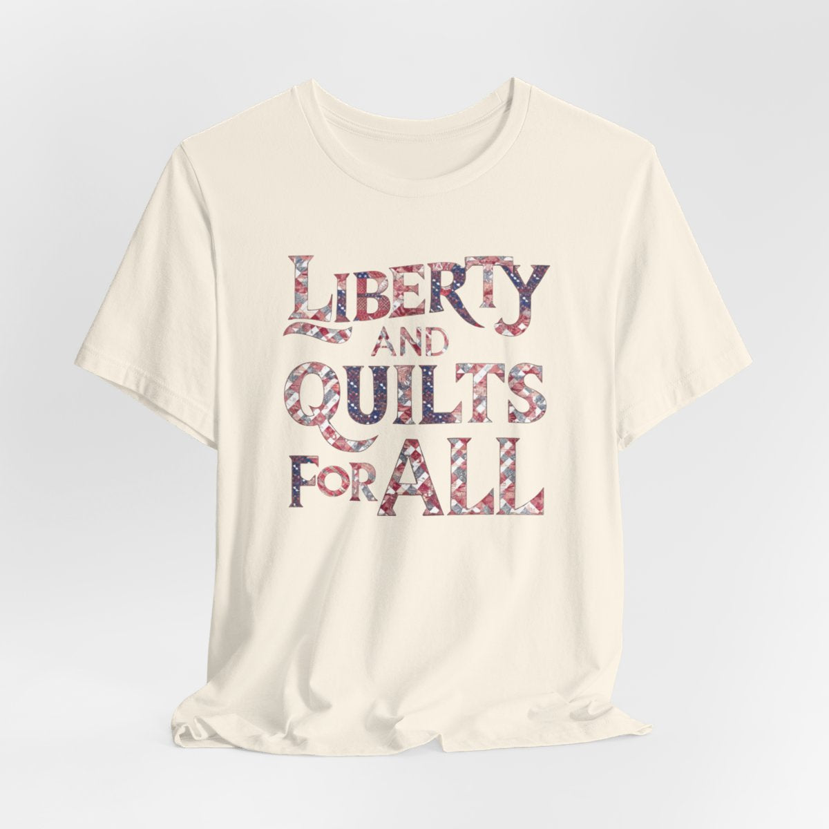 A Natural T-shirt featuring text "LIBERTY AND QUILTS FOR ALL" in large decorative letters filled with various quilt patterns in red, white, and blue.