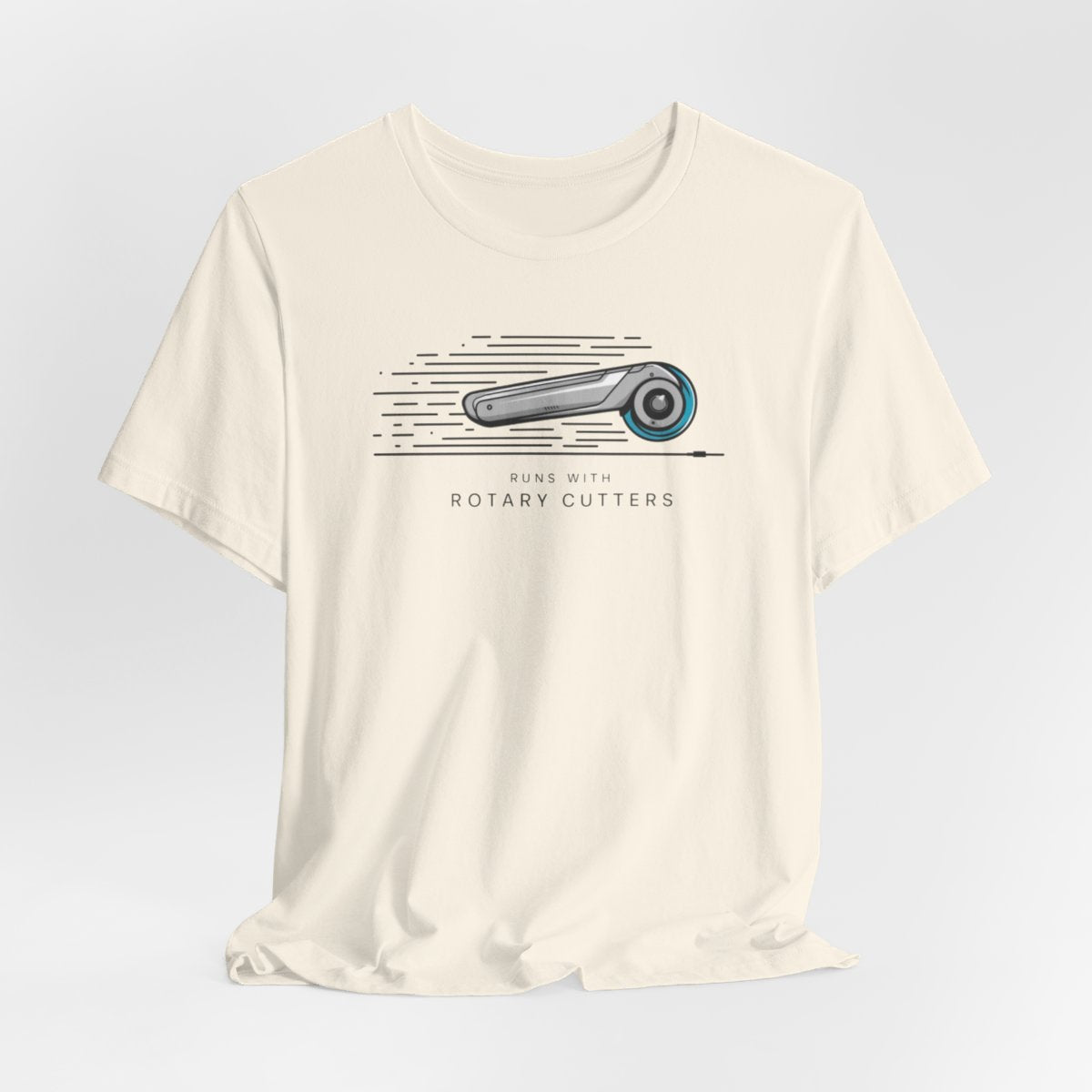 A Natural T-shirt featuring minimalist graphic of a sleek rotary cutter in motion with speed lines. Text below reads "RUNS WITH ROTARY CUTTERS" in a modern font.