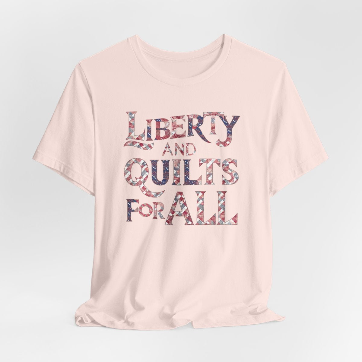 A Soft Pink T-shirt featuring text "LIBERTY AND QUILTS FOR ALL" in large decorative letters filled with various quilt patterns in red, white, and blue.